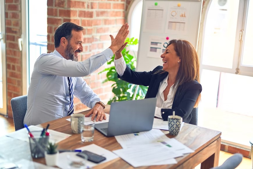 man and woman in professional attire sitting at a table together and high-fiving