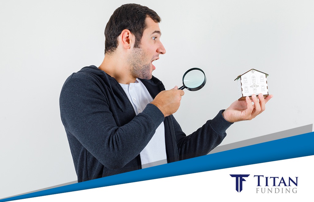 Man in a sweater holding a magnifying glass in one hand and a miniature house in the other