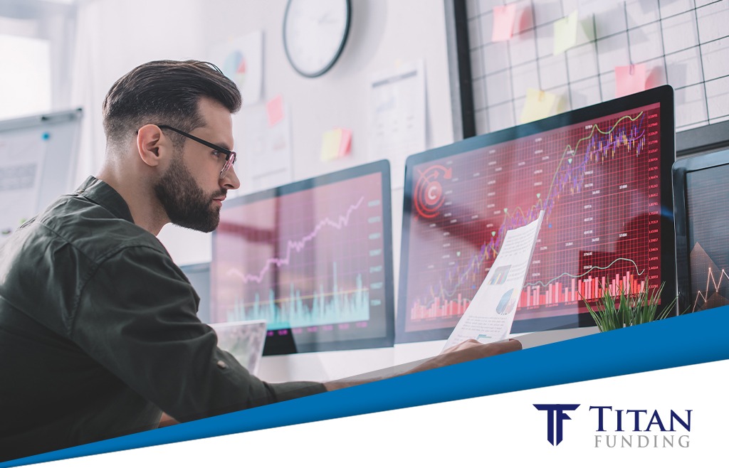 Man with glasses looking at a financial document while sitting at a desk with two computer screens